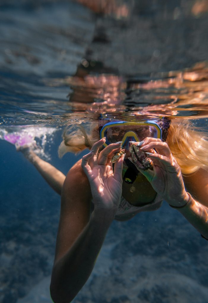 Unrecognizable woman in swimwear and snorkeling mask with flippers showing seashells while swimming underwater near reef in clear blue sea